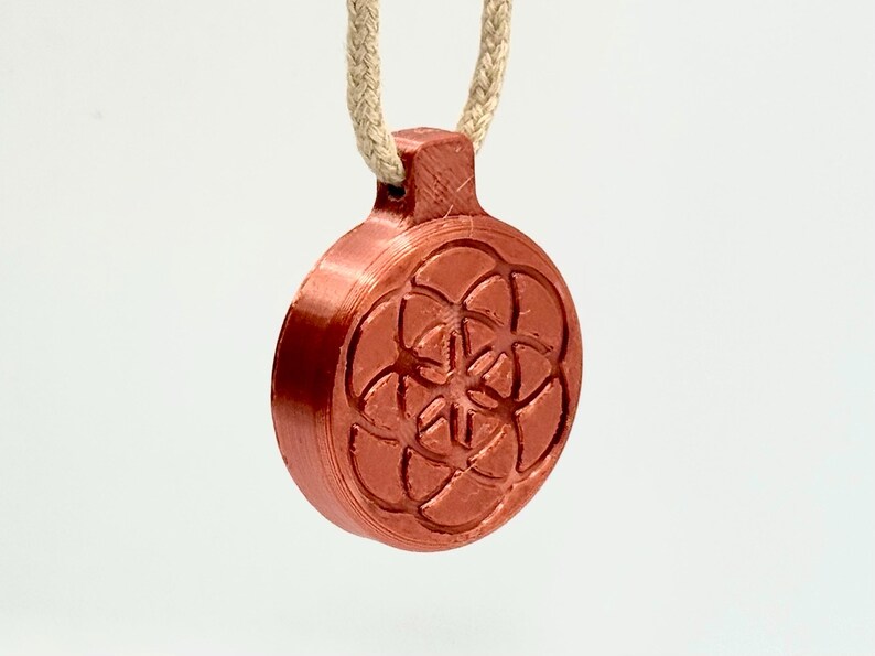 Op de afbeelding: Een koperkleurige hanger met een geometrisch bloem des levens-ontwerp, hangend aan een beige touw. De ronde hanger heeft een diameter van 3,8 cm en heeft een gedetailleerd, verhoogd patroon. Het touw is ongeveer 25 cm lang.