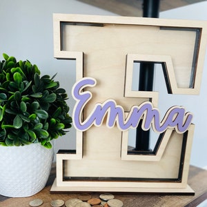 May include: A wooden piggy bank shaped like the letter E with the name 'Emma' written in purple cursive on the front. The piggy bank is sitting on a wooden surface with a pile of coins in front of it.