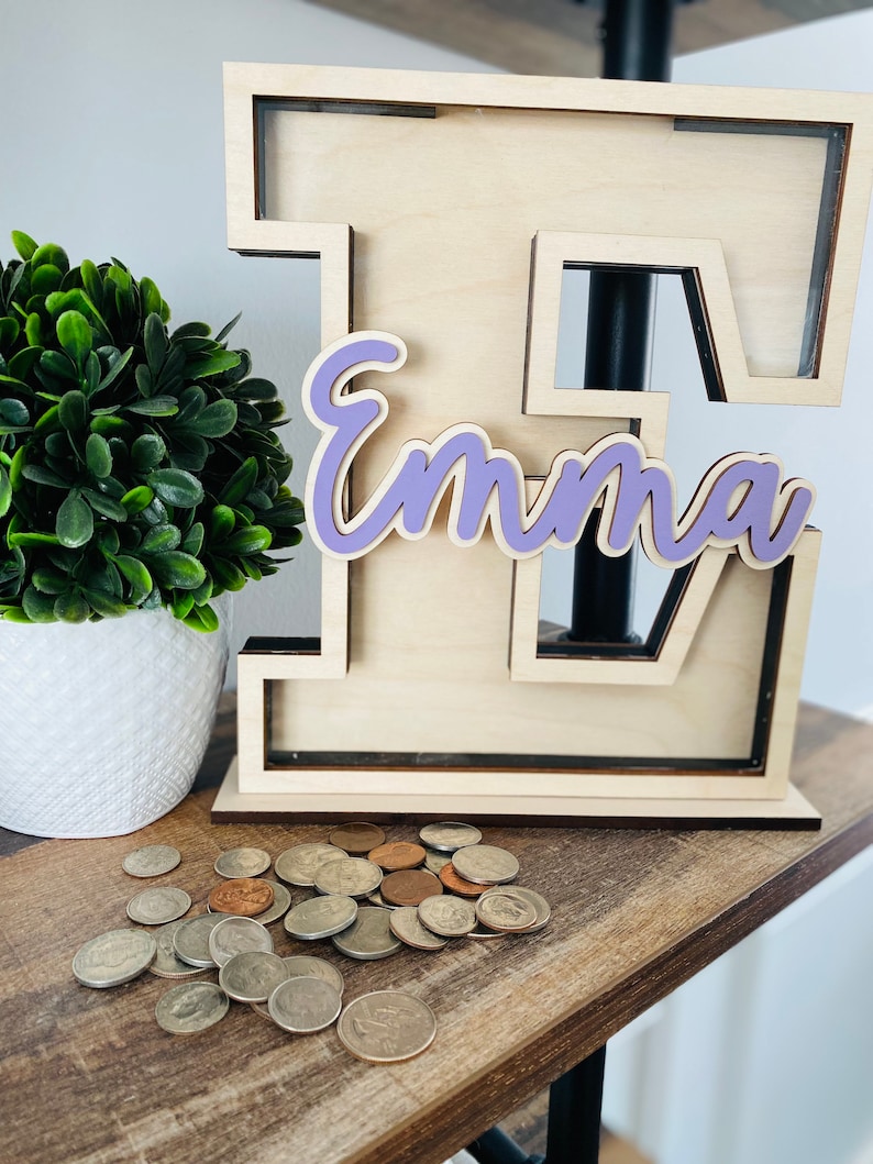 May include: A wooden piggy bank shaped like the letter E with the name 'Emma' written in purple cursive on the front. The piggy bank is sitting on a wooden surface with a pile of coins in front of it.