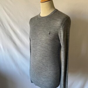 May include: A gray crew neck sweater with long sleeves. The sweater has a small, dark emblem on the left chest. The garment is displayed on a wooden mannequin against a white backdrop. The sweater is made of a soft, knit material.