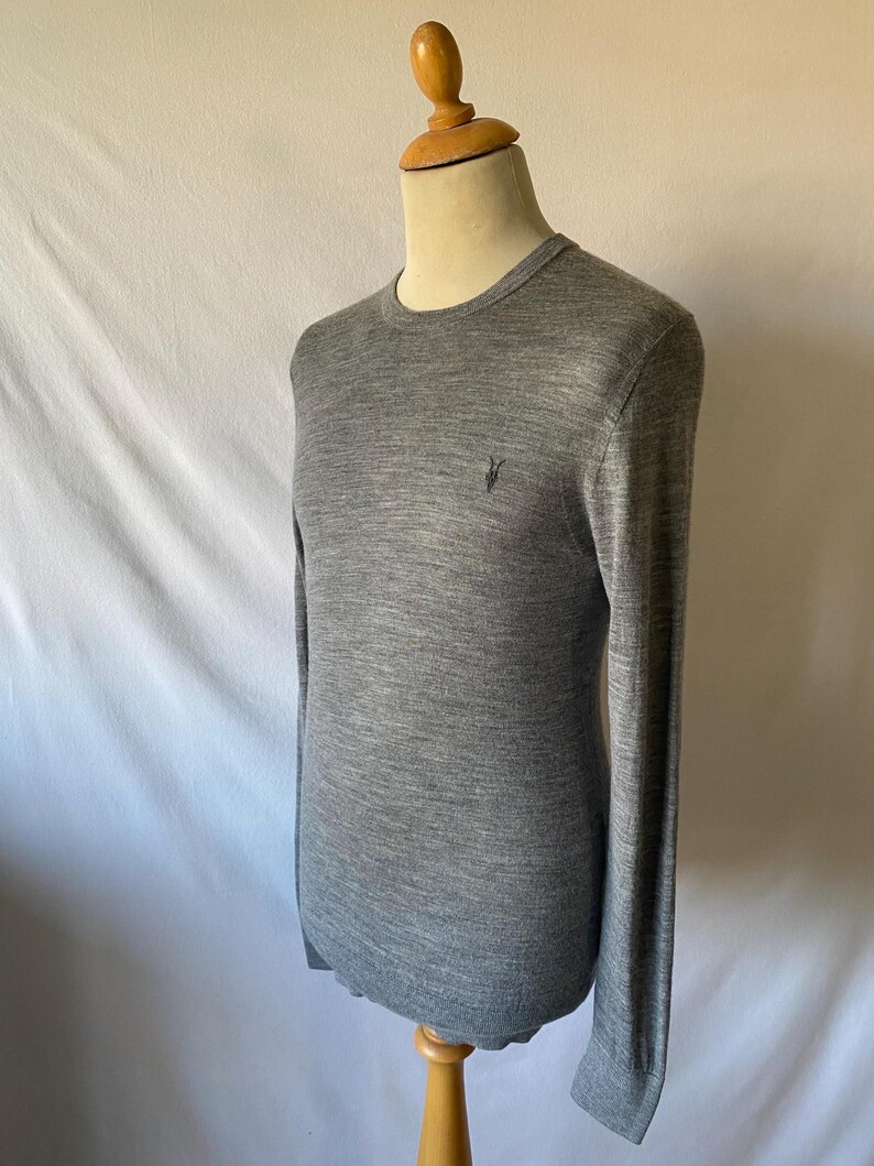 May include: A gray crew neck sweater with long sleeves. The sweater has a small, dark emblem on the left chest. The garment is displayed on a wooden mannequin against a white backdrop. The sweater is made of a soft, knit material.