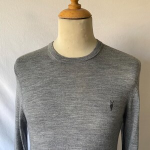 May include: A heather gray long-sleeve sweater with a crew neck. The sweater has a small black emblem on the chest. The garment is displayed on a mannequin.
