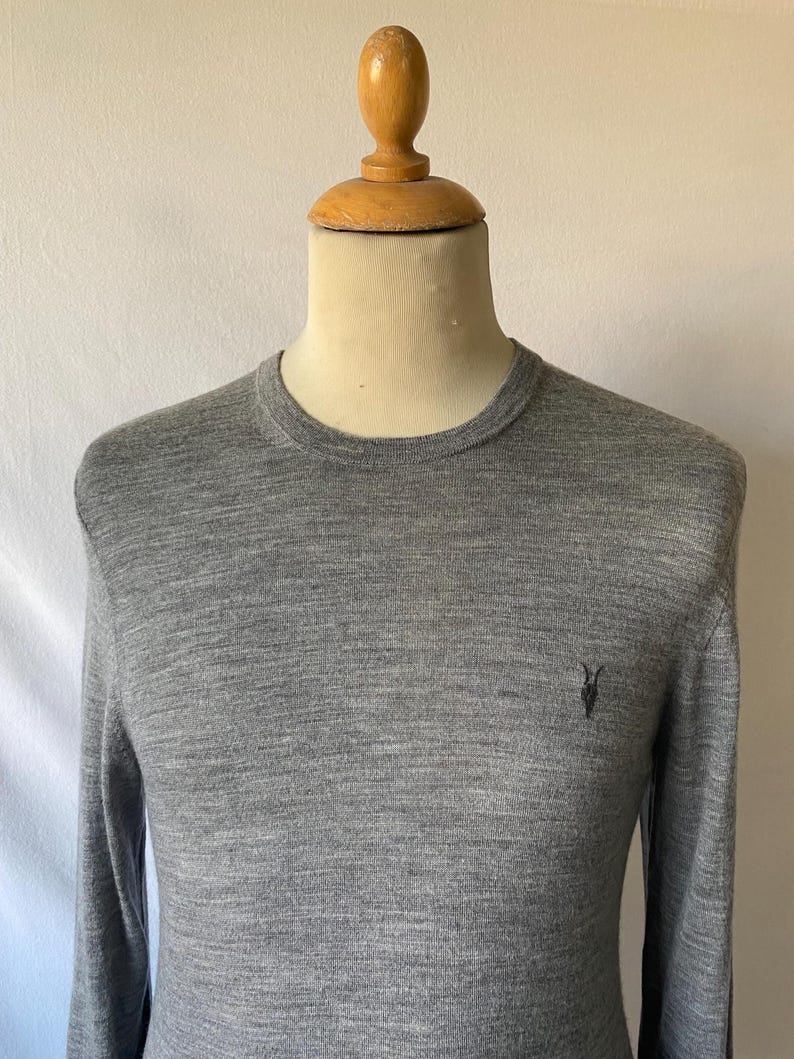 May include: A heather gray long-sleeve sweater with a crew neck. The sweater has a small black emblem on the chest. The garment is displayed on a mannequin.