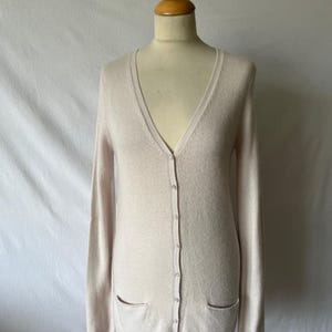 K&ouml;nnte beinhalten: Ein hellbeige Cardigan mit V-Ausschnitt, langen &Auml;rmeln und zwei Vordertaschen. Der Cardigan ist zugekn&ouml;pft und auf einer Schaufensterpuppe ausgestellt. Der Stoff wirkt weich und leicht, geeignet zum &Uuml;berziehen. Der Gesamtstil ist klassisch und vielseitig.