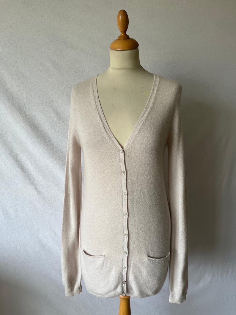K&ouml;nnte beinhalten: Ein hellbeige Cardigan mit V-Ausschnitt, langen &Auml;rmeln und zwei Vordertaschen. Der Cardigan ist zugekn&ouml;pft und auf einer Schaufensterpuppe ausgestellt. Der Stoff wirkt weich und leicht, geeignet zum &Uuml;berziehen. Der Gesamtstil ist klassisch und vielseitig.