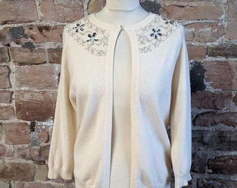 May include: A cream-coloured cardigan with a beaded neckline featuring a floral design and clear rhinestones. The cardigan has a front opening and long sleeves.