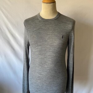 May include: A heather gray long-sleeve sweater with a crew neck, displayed on a mannequin. The sweater has a small, dark emblem on the chest. The fabric appears to be a fine knit, suitable for layering or wearing on its own.