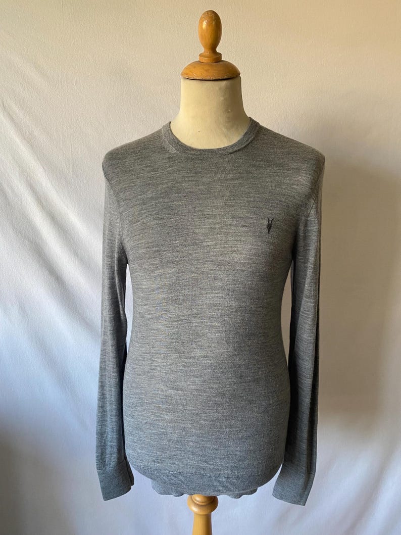 May include: A heather gray long-sleeve sweater with a crew neck, displayed on a mannequin. The sweater has a small, dark emblem on the chest. The fabric appears to be a fine knit, suitable for layering or wearing on its own.