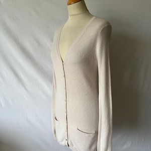 K&ouml;nnte beinhalten: Ein hellbeige Cardigan mit V-Ausschnitt und langen &Auml;rmeln. Der Cardigan hat zwei Vordertaschen und einen Knopfverschluss. Er wird auf einer Schaufensterpuppe vor wei&szlig;em Hintergrund pr&auml;sentiert. Das Kleidungsst&uuml;ck ist aus weichem Strickmaterial.