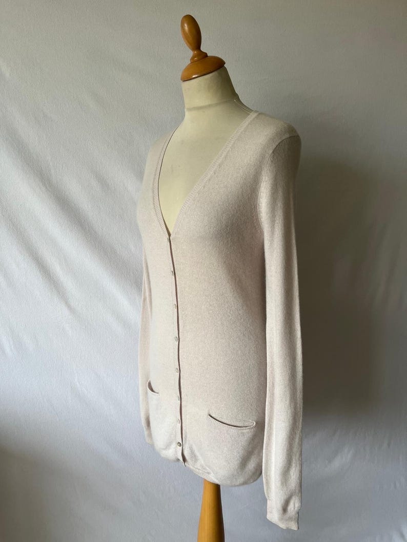 K&ouml;nnte beinhalten: Ein hellbeige Cardigan mit V-Ausschnitt und langen &Auml;rmeln. Der Cardigan hat zwei Vordertaschen und einen Knopfverschluss. Er wird auf einer Schaufensterpuppe vor wei&szlig;em Hintergrund pr&auml;sentiert. Das Kleidungsst&uuml;ck ist aus weichem Strickmaterial.