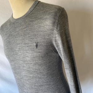 May include: A heather gray long-sleeve sweater with a crew neck. The sweater features a small, dark embroidered emblem on the chest. The fabric appears to be a fine knit, and the sleeves are long.