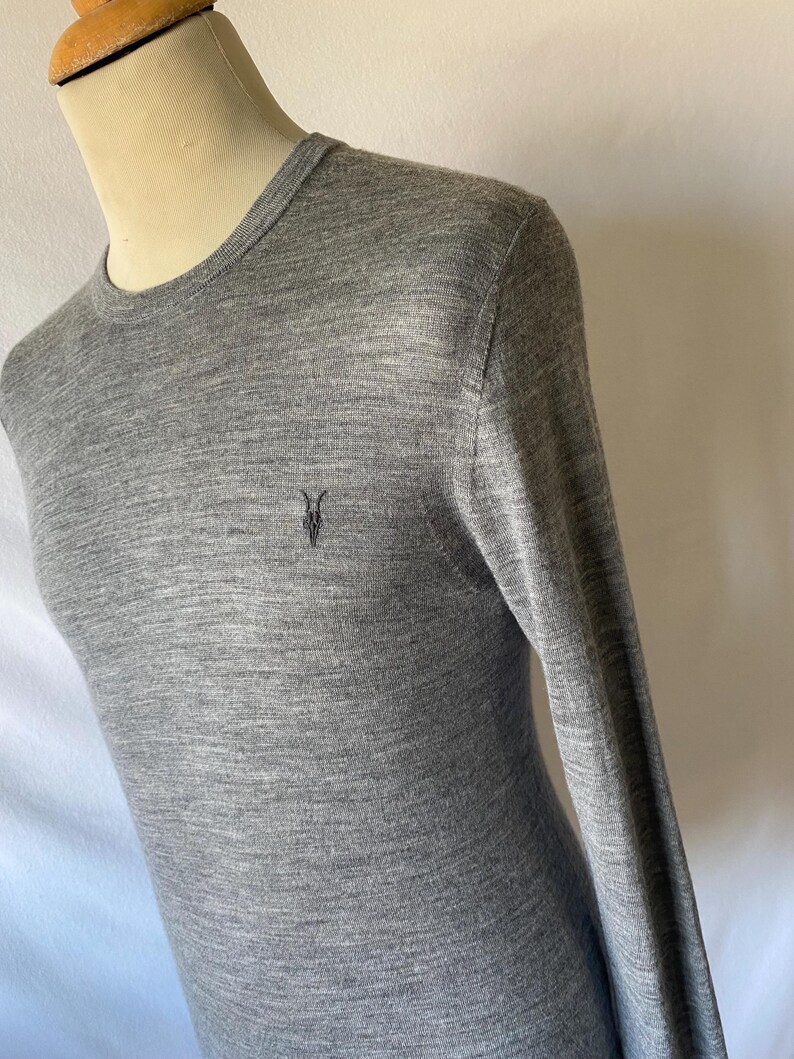 May include: A heather gray long-sleeve sweater with a crew neck. The sweater features a small, dark embroidered emblem on the chest. The fabric appears to be a fine knit, and the sleeves are long.
