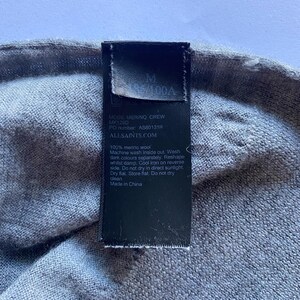 May include: Close-up of a gray merino wool garment with a black tag. The tag displays the size "M" and the text "MODE MERINO CREW" along with care instructions and the brand's website.