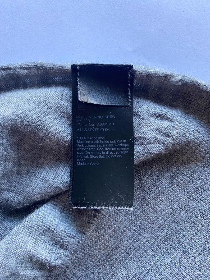 May include: Close-up of a gray merino wool garment with a black tag. The tag displays the size "M" and the text "MODE MERINO CREW" along with care instructions and the brand's website.