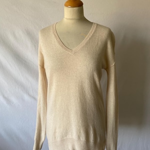 Può includere: Un maglione beige chiaro con scollo a V esposto su un manichino. Il maglione ha maniche lunghe e dettagli a coste sull'orlo, sui polsini e sullo scollo. Il tessuto sembra morbido e leggero, con uno stile semplice e classico.