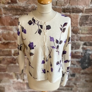 Peut inclure: Un cardigan crème avec un motif floral dans les tons de violet et de marron. Le cardigan a une bordure en crochet et une fermeture à boutons.