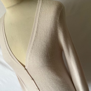 K&ouml;nnte beinhalten: Hellbeige Strickjacke mit V-Ausschnitt und langen &Auml;rmeln. Der Strickpullover hat eine Knopfleiste vorne. Das Kleidungsst&uuml;ck wird auf einer Schaufensterpuppe pr&auml;sentiert.