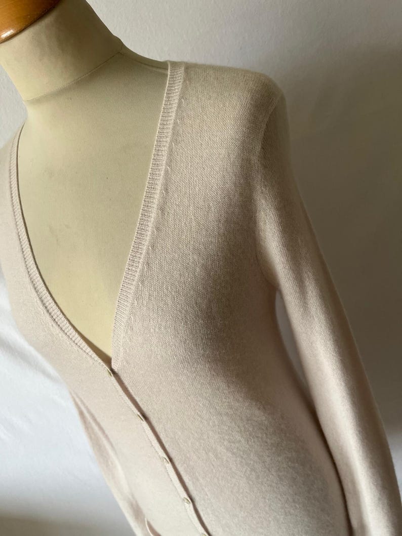 K&ouml;nnte beinhalten: Hellbeige Strickjacke mit V-Ausschnitt und langen &Auml;rmeln. Der Strickpullover hat eine Knopfleiste vorne. Das Kleidungsst&uuml;ck wird auf einer Schaufensterpuppe pr&auml;sentiert.