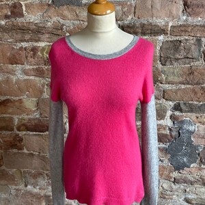 Può includere: Un maglione di cashmere rosa con bordino grigio intorno al collo e maniche grigie.