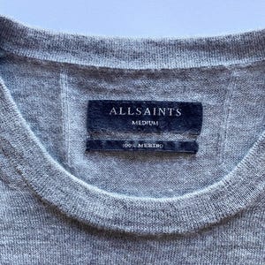 May include: Close-up of a gray crew neck sweater. The label reads "ALLSAINTS MEDIUM 100% MERINO". The sweater is made of a soft, knitted material. The garment has a ribbed neckline.