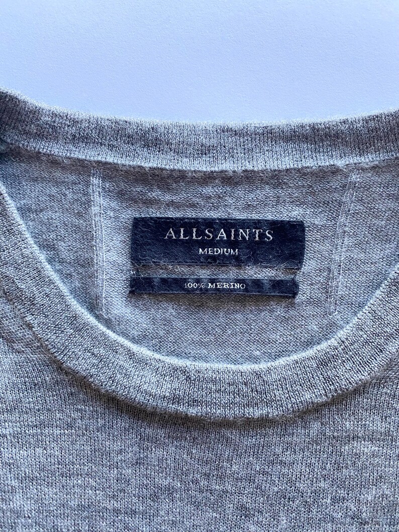 May include: Close-up of a gray crew neck sweater. The label reads "ALLSAINTS MEDIUM 100% MERINO". The sweater is made of a soft, knitted material. The garment has a ribbed neckline.