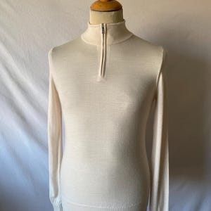 May include: A light beige long-sleeve sweater with a mock neck and a quarter-zip closure. The sweater is displayed on a wooden mannequin. The fabric appears to be a fine knit.
