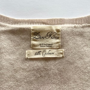 K&ouml;nnte beinhalten: Nahaufnahme eines hellbeigen Zara Knit-Pullovers. Das Etikett lautet "Zara Knit" und "100% Cashmere". Der Pullover wird in Thailand hergestellt. Der Pullover hat eine neutrale Farbe.