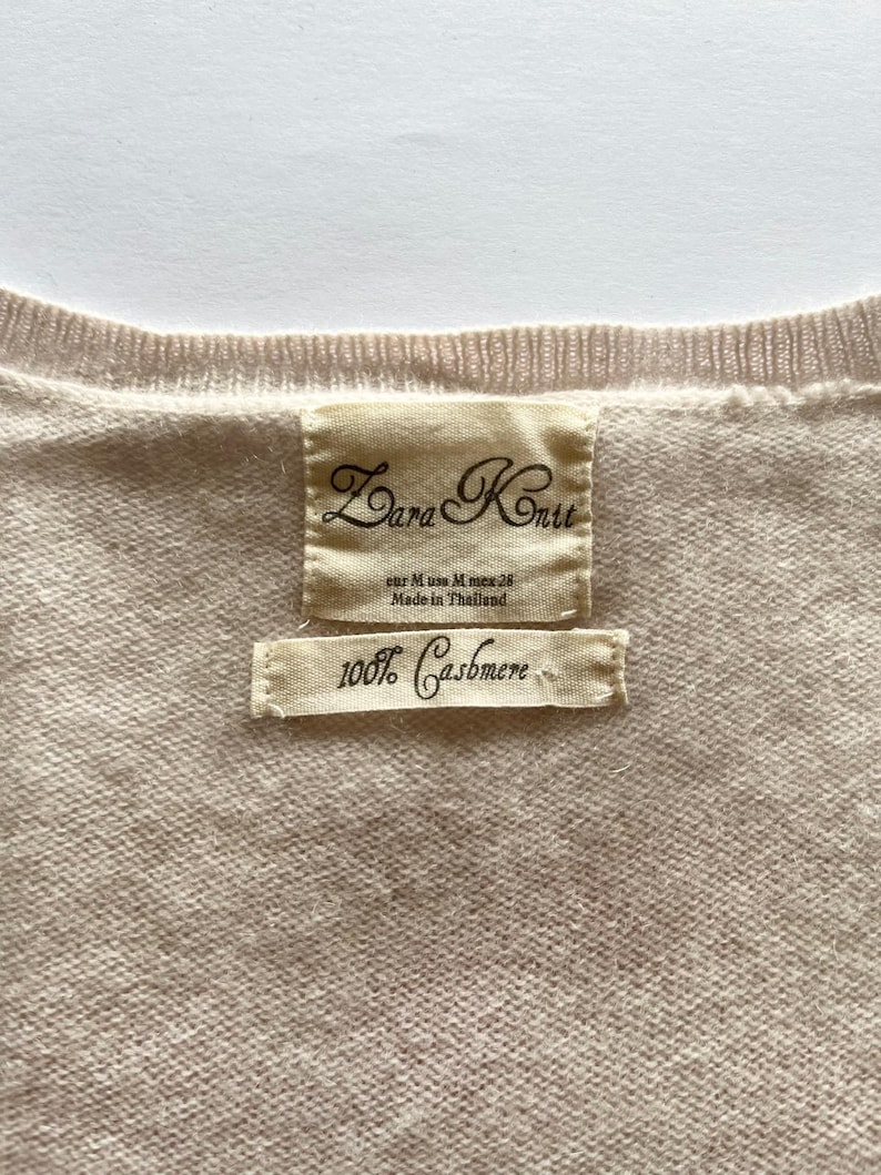 K&ouml;nnte beinhalten: Nahaufnahme eines hellbeigen Zara Knit-Pullovers. Das Etikett lautet "Zara Knit" und "100% Cashmere". Der Pullover wird in Thailand hergestellt. Der Pullover hat eine neutrale Farbe.