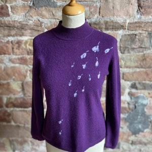 May include: A purple long-sleeved sweater with a high neck and a delicate floral design on the right shoulder.