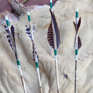 May include: Four brown wooden arrows with green and white accents and fletching made of feathers. The arrows are lying on a tan, textured surface.
