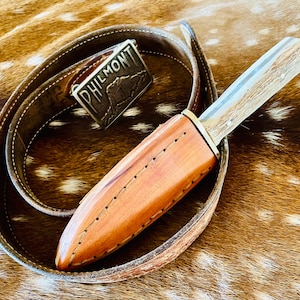 May include: A handcrafted hunting knife with a light-colored handle and a leather sheath. The knife is resting on a brown leather belt with a silver buckle that reads "PHILMONT". The belt is wrapped around the knife and sheath.