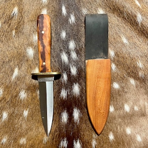 Könnte beinhalten: Ein kleines Messer mit braunem Griff, silberner Klinge und einer Lederscheide. Das Messer liegt auf einer braunen und weiß gefleckten Oberfläche.