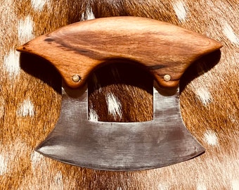 May include: A stainless steel mezzaluna knife with a curved blade and a wooden handle. The handle is made of light brown wood and is attached to the blade with two small metal rivets. The knife is designed for chopping herbs and vegetables.