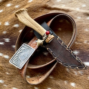 May include: A brown leather belt with a silver buckle that says "PHILMONT" and a knife with a bone handle and a brown leather sheath.