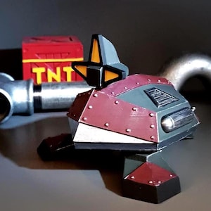 May include: A grey and red robot toy with a yellow and black antenna. The robot has a silver panel on the front and a black cylinder on the side. The robot is sitting on a grey surface. The word "TNT" is visible on a red box in the background.