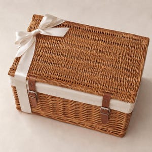A closed brown woven wicker basket with a white liner, two brown leather straps with metal buckles, and a white satin ribbon tied in a bow on top.