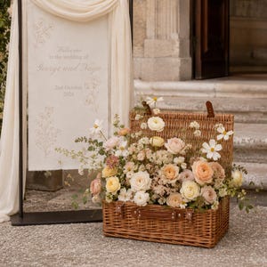 Large wicker wedding hamper filled with romantic flowers beside a wedding welcome sign, styled as rustic wedding décor for an outdoor venue entrance.