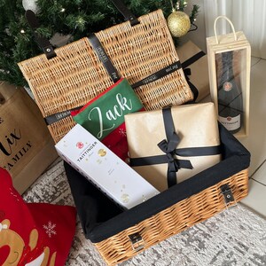 Op de afbeelding: Een kerstcadeaumand gevuld met cadeaus, een fles champagne in een houten kist en een groene pouch met de naam "Jack". De mand is gemaakt van geweven riet en heeft zwarte handvatten en een zwart interieur.