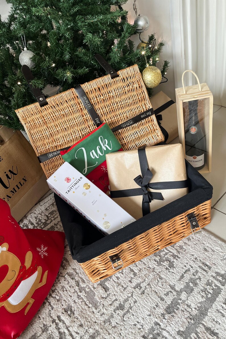 Op de afbeelding: Een kerstcadeaumand gevuld met cadeaus, een fles champagne in een houten kist en een groene pouch met de naam "Jack". De mand is gemaakt van geweven riet en heeft zwarte handvatten en een zwart interieur.