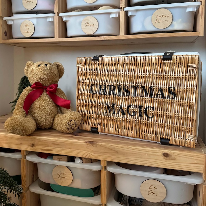 May include: A brown teddy bear with a red ribbon sits next to a woven wicker basket. The basket has the words "CHRISTMAS MAGIC" in black lettering. The scene is set on a wooden shelf with storage bins.