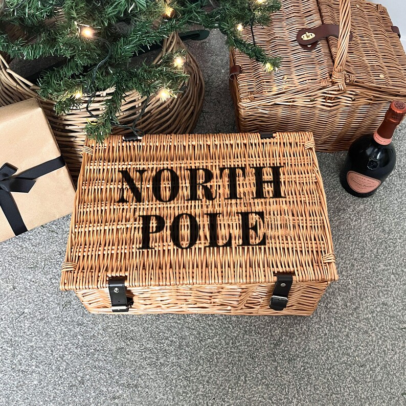 Op de afbeelding: Wicker picknickmand met de woorden "NORTH POLE" in zwarte letters. De mand is rechthoekig met een scharnierend deksel en leren riemen. Een kerstboom, een geschenkdoos en een fles wijn staan op de achtergrond.
