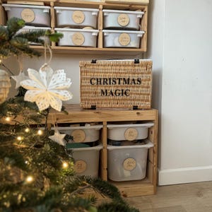 Op de afbeelding: Een houten opbergunit met meerdere witte bakken en een gevlochten mand met het opschrift "CHRISTMAS MAGIC". De bakken hebben houten labels. Een kerstboom met lichtjes en versieringen staat op de voorgrond.