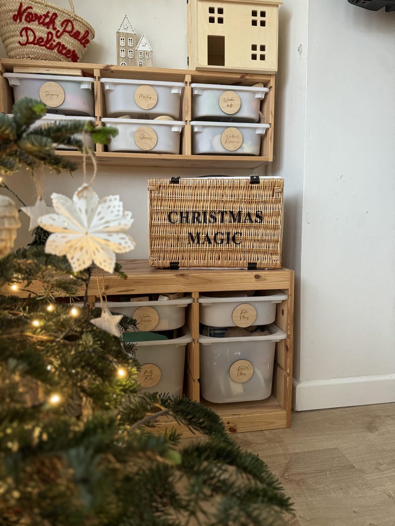 Op de afbeelding: Een houten opbergunit met meerdere witte bakken en een gevlochten mand met het opschrift "CHRISTMAS MAGIC". De bakken hebben houten labels. Een kerstboom met lichtjes en versieringen staat op de voorgrond.