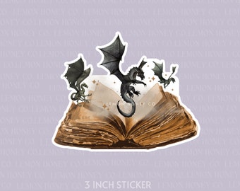 May include: A sticker with a fantasy theme. It shows an open book with three black dragons flying above it. The sticker is approximately 7.6 cm in size and has the text "Lemon Honey Co."