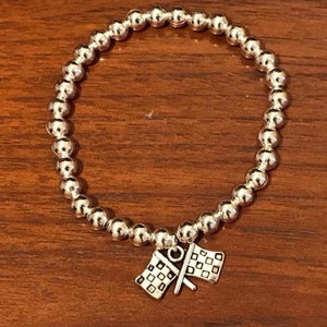 Könnte beinhalten: Ein kreisförmiges Stretch-Armband aus kleinen, polierten silberfarbenen Metallperlen. Am unteren Teil des Armbands befindet sich ein Anhänger, der zwei gekreuzte karierte Rennflaggen mit einem detaillierten Gittermuster aus silbernem Metall zeigt.