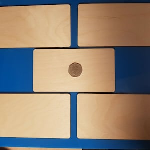Op de afbeelding: Vijf lege rechthoekige houten plaquettes op een blauwe achtergrond. De plaquettes zijn gerangschikt in een rasterpatroon met één plaquette in het midden en twee plaquettes erboven en eronder. Een munt is op de middelste plaquette geplaatst.