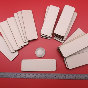 Pode incluir: Uma pilha de 20 peças retangulares de madeira inacabadas, com aproximadamente 7,6 cm de comprimento e 2,5 cm de largura, sobre um fundo vermelho. Uma única peça é colocada na frente da pilha, e uma moeda de prata é colocada ao lado.