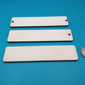 May include: Three unfinished wooden rectangular signs with rounded edges and holes in the top corners. The signs are stacked on top of each other on a blue surface.