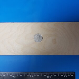 May include: A rectangular, light-coloured wooden board with a coin on top. The board is approximately 30 cm long. A ruler is at the bottom of the image, with measurements in centimetres.