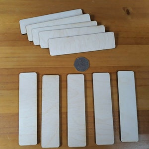 Könnte beinhalten: Sieben unfertige rechteckige Holzplaketten, perfekt zum Basteln und Personalisieren. Die Plaketten sind fächerförmig angeordnet, wobei die größte Plakette unten liegt. Eine kleine Münze liegt vor den Plaketten.